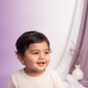 Close-up portrait of a smiling South Asian toddler with a curious and thoughtful expression, against a soft lavender and whit